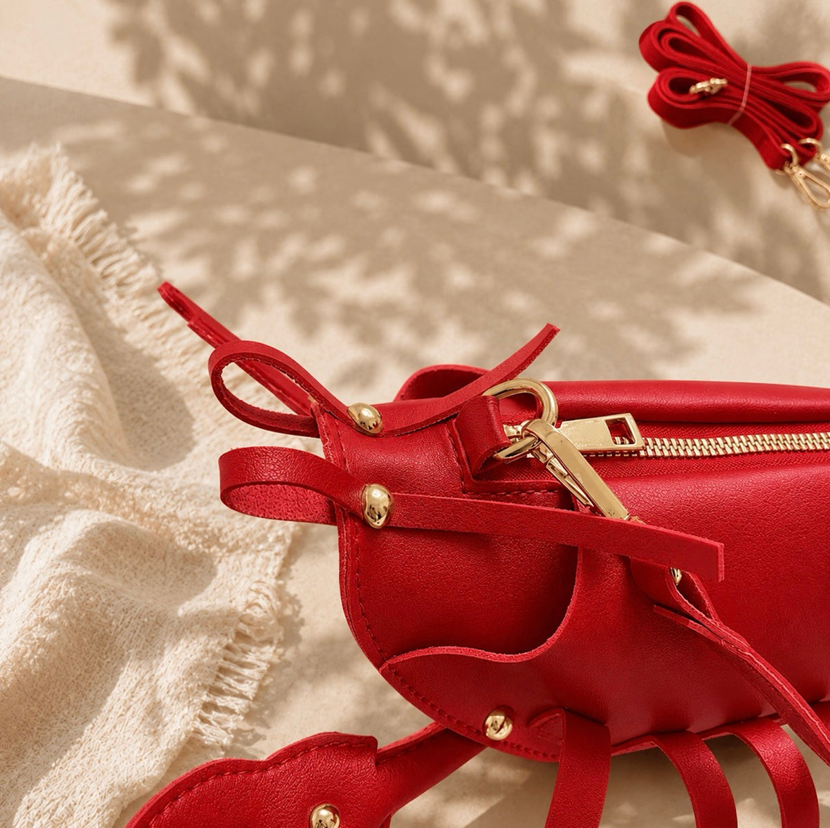 A close-up of a bright red leather bag with decorative tie straps, gold-tone hardware, and a gold zipper, styled on a beige textured surface with a cream knit blanket and leaf shadows.