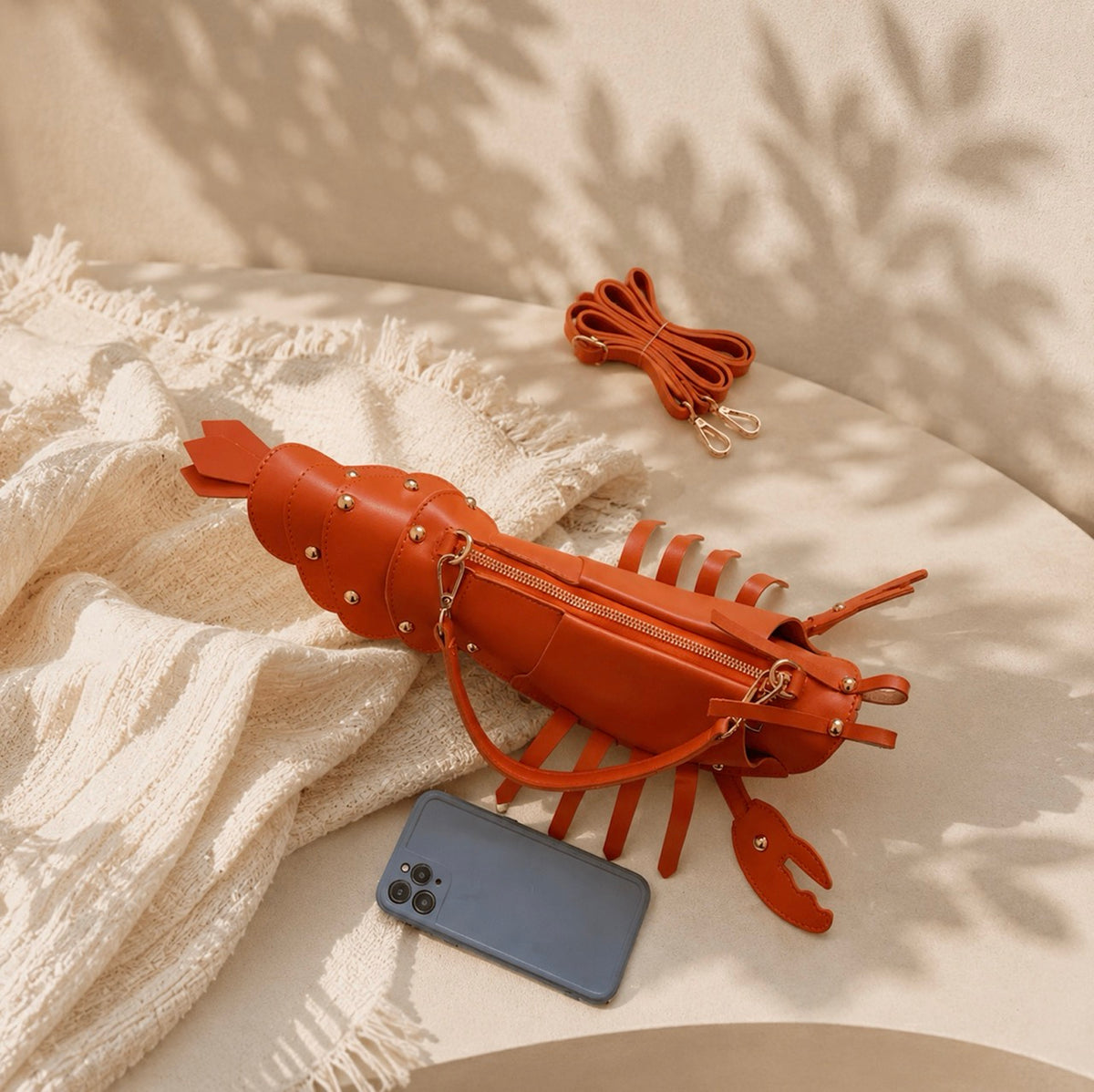 An orange lobster-shaped crossbody bag, styled on a cream textured blanket with a smartphone and its strap nearby, under soft leaf shadows.