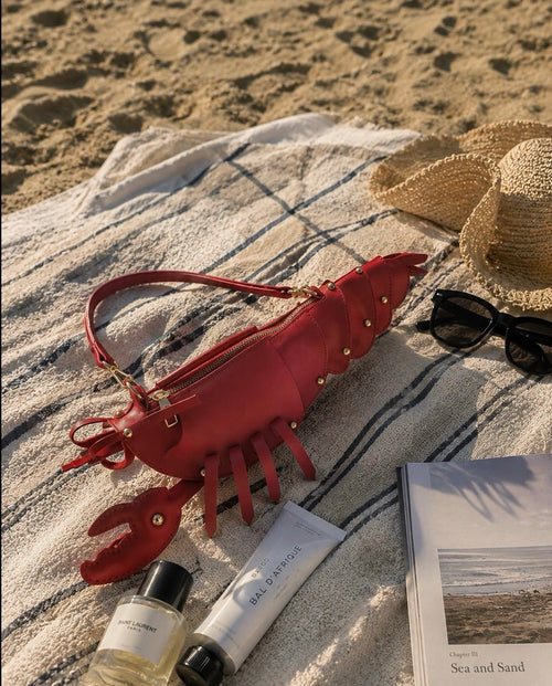 A lobster-shaped red leather handbag on a beach towel with a straw hat, black sunglasses, a perfume bottle, and a tube of lip balm on a sandy beach.