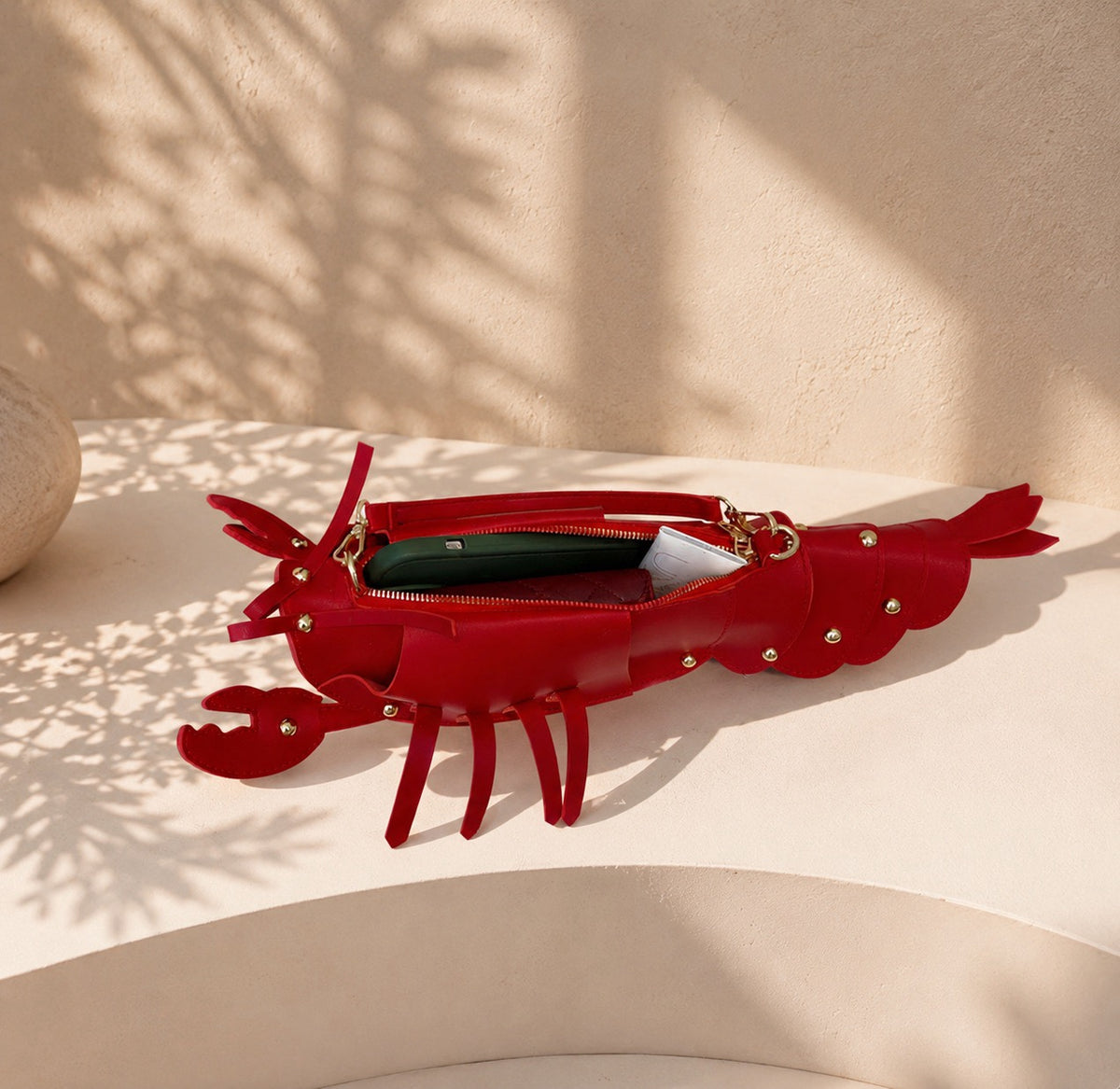 A red lobster-shaped zip pouch bag with gold-tone studs and hardware, shown open with mobile phone, purse and other items inside - on a beige platform with dappled shadows.